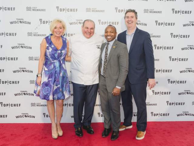 Four people stand smiling on a red carpet in front of a backdrop with Top Chef, Visit Milwaukee, and Travel Wisconsin logos. They are dressed in formal and business attire and pose together for a photo.
