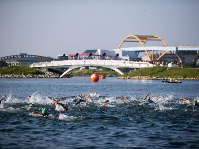 A group of swimmers wearing green caps race through open water during a triathlon, with splashing waves in the foreground and spectators watching from a bridge near Milwaukee’s Hoan Bridge in the background.