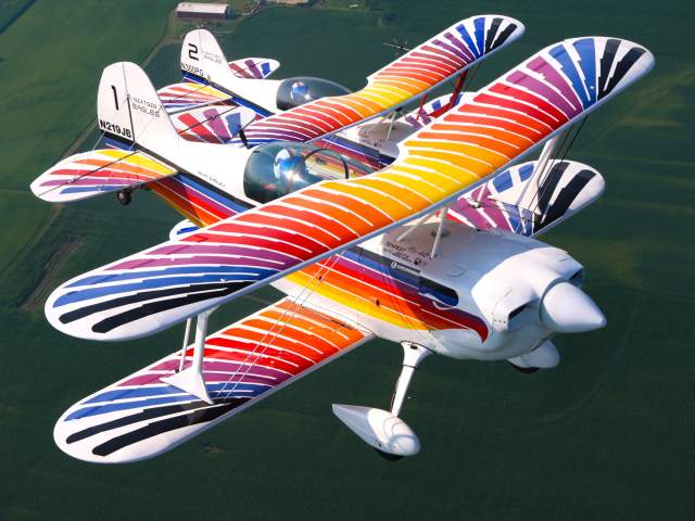 Two brightly colored biplanes fly in close formation above green water, showcasing coordinated aerobatic precision. Their vibrant red, orange, blue, and white striped wings create a striking visual display against the calm background below.