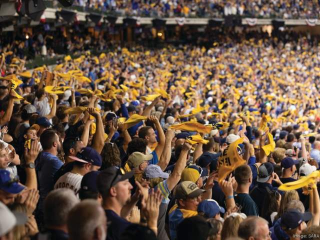 A packed stadium crowd cheers and waves yellow rally towels during a baseball game, filling the stands with motion and team colors.