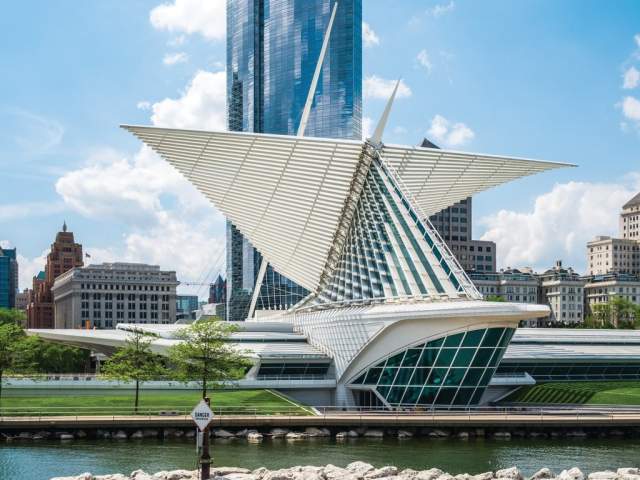 A daytime view of the Milwaukee Art Museum with its white, wing-like brise soleil extended against a blue sky. The sleek glass-and-steel structure sits along the lakefront, with a rocky shoreline in the foreground and tall downtown buildings rising behind it.