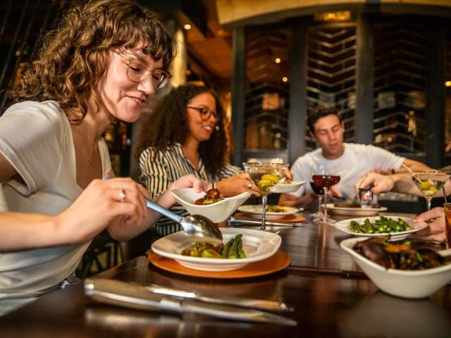 A group of four people sit together at a restaurant table, sharing small plates of food. A woman in the foreground smiles as she serves herself from a bowl, while others reach for dishes and enjoy cocktails. The setting is warm and inviting, with dim lighting and a stylish interior.