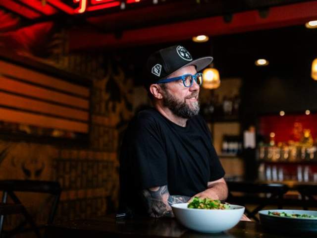 A man wearing a black T-shirt, black cap, and blue glasses sits at a restaurant table with bowls of food in front of him. Warm hanging lights and colorful wall art create a cozy, modern atmosphere in the background.