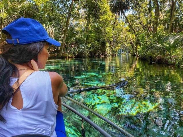 A woman in a tank top and baseball cap sits in a clear kayak, admiring clear aqua water at Rainbow Springs State Paek