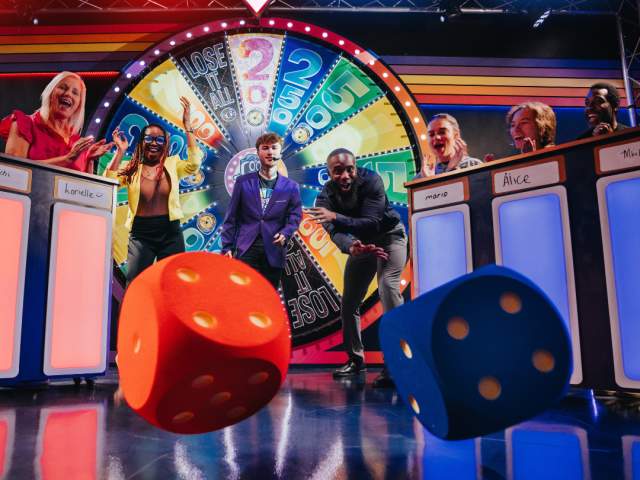 Contestants cheer as they compete in a game show-inspired attraction. Two contestants throw giant dice as their friends look on, a giant spinning wheel of luck in the background.