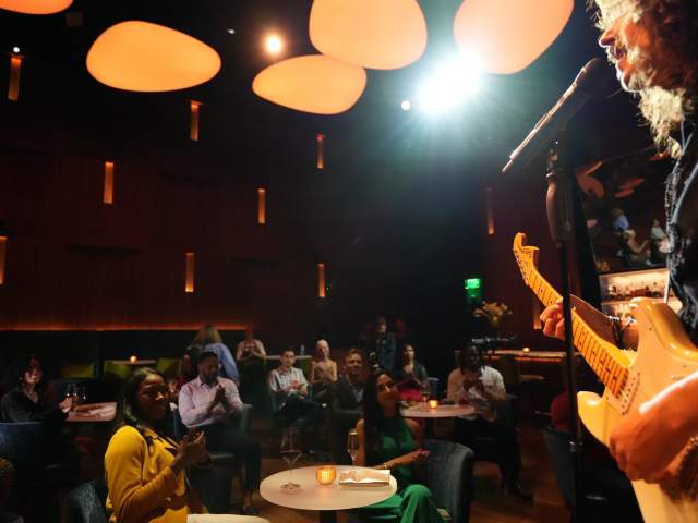 A singer holding a guitar stands to the right of the frame, looking out at an audience seated at intimate listening lounge tables