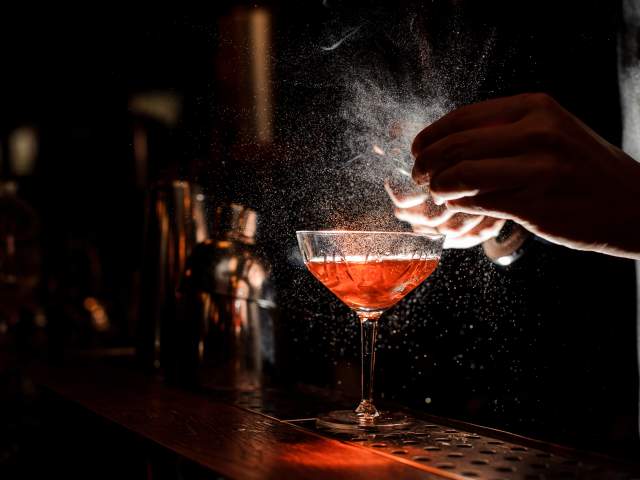 Barmans hands sprinkling the juice into the cocktail glass filled with alcoholic drink on the dark background