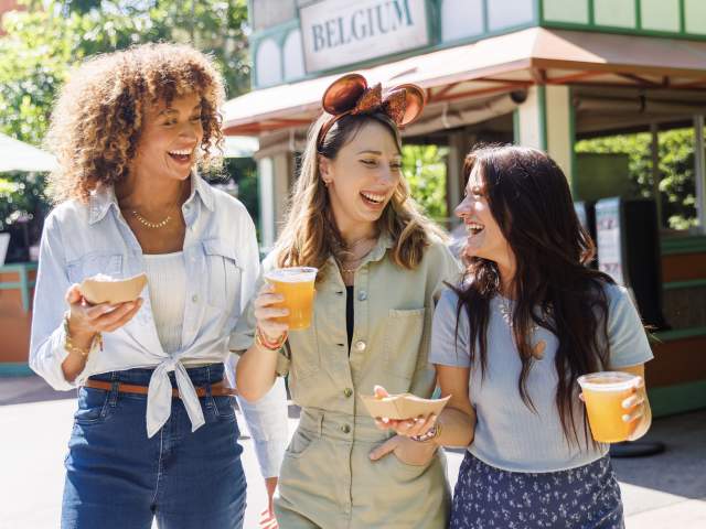Three women walking at EPCOT enjoying drinks and food from a festival booth.