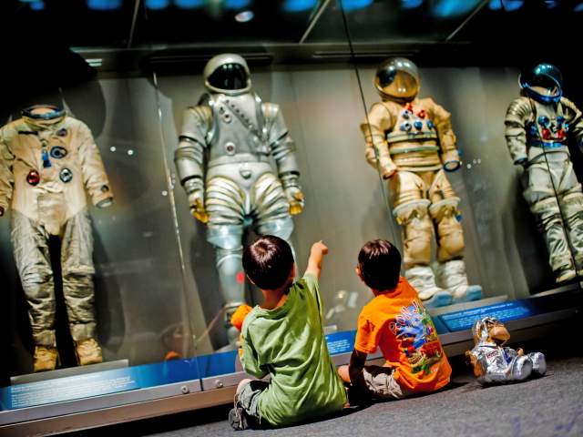 Two children point and gaze up at astronaut spacesuits in the Kennedy Space Center Visitor Complex treasures gallery
