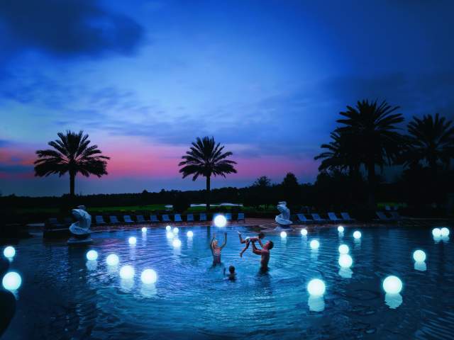 A family plays with glowing beach balls at the pool as day turns to night