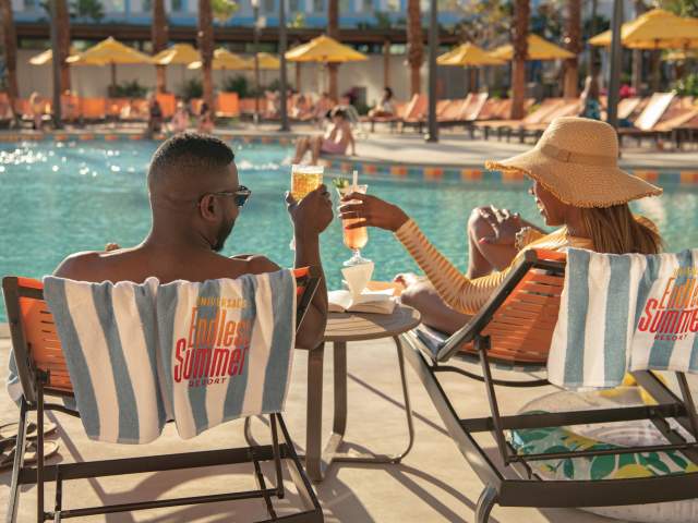 A man and a woman unwind by the pool at Universal Endless Summer Resort – Dockside Inn and Suites. They are seen from behind, raising celebratory cocktails while overlooking the sparkling pool.