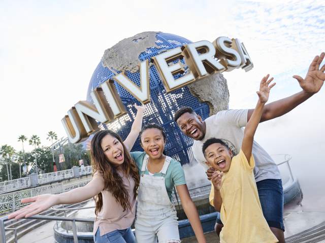 A smiling family of four — mom, dad, preteen daughter and preteen son — stand in front of the Universal globe, their arms outstretched and welcoming.