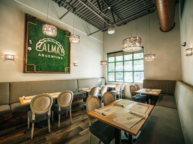 Interior dining room at Alma Argentina featuring booth seating, upholstered chairs, decorative lighting, branded wall signage, and neatly arranged dining tables.