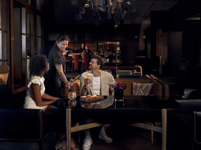A hospitality team member serves beverages to guests seated in the lounge area at Capa. Contemporary furnishings, specialty cocktails, and personalized service reflect the restaurant's upscale atmosphere and guest-focused dining experience.