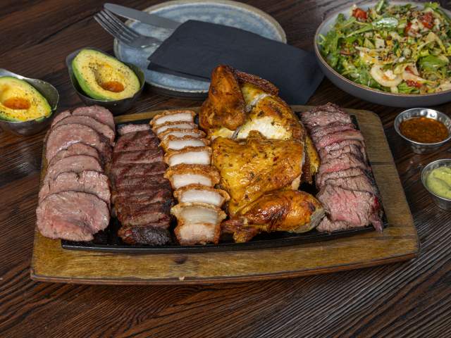 A large platter features assorted grilled meats including chicken, steak, and sliced pork, accompanied by avocado halves, sauces, and salad. The dish is arranged for shared dining.