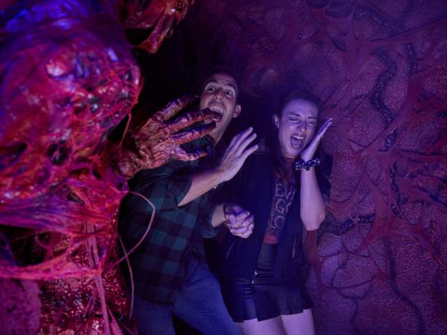 Two young adults flinch and scream in terror, recoiling as a monstrous red creature reaches for them at Halloween Horror Nights