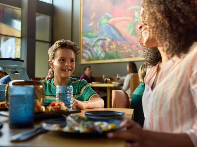 A son looks at his mother and smiles over a meal at BACAN, the restaurant's signature mural prominent in the background
