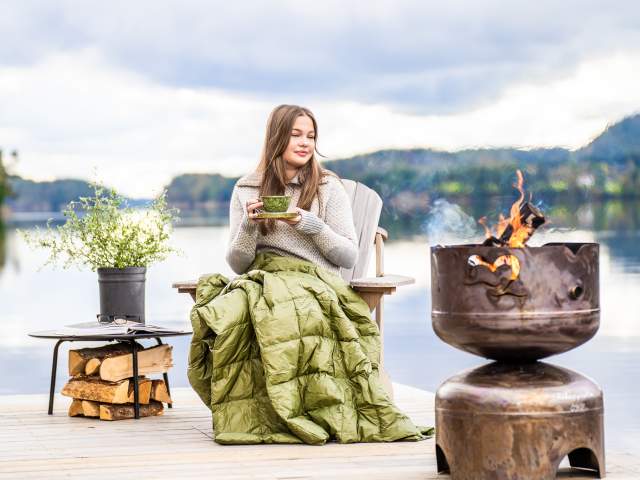 Ung kvinne nyter en kaffekopp ved bålpanne på FLO Haldenkanalen med utsikt over innsjø.