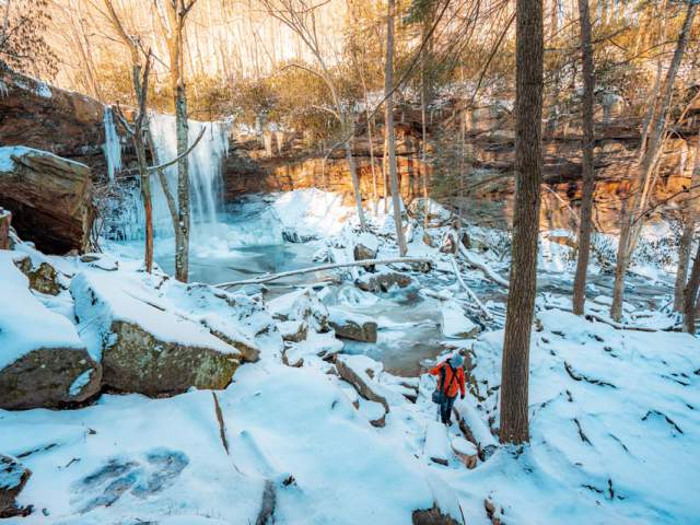 Person in snow gear walking across snow covered ground in the woods with the waterfall Ohiopyle Cucumber Falls in the background.