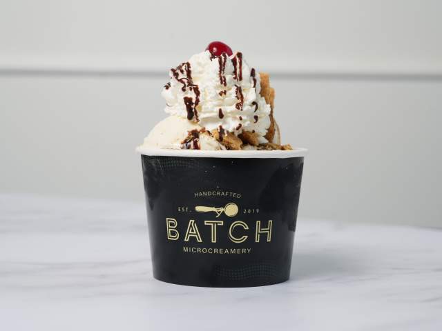 A cup of ice cream with whipped cream, chocolate sauce, and a cherry on top in a cup that reads Batch