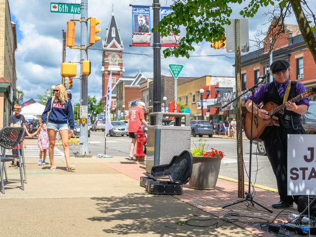 A woman holding a young child's hand while walking down a main street sidewalk with a man playing a guitar