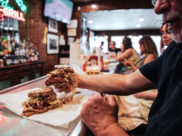 Man sitting at a bar counter holding up a half of a sandwich that has meat, cheese, and french fries piled up