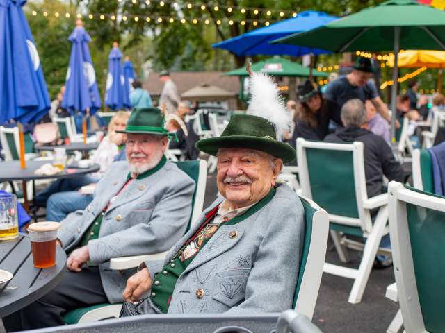 Two older men in green hats sitting in chairs at an outdoor restaurant with pints of beer in front of them
