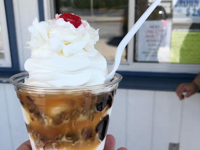 A person holding up an ice cream cup with caramel, whipped cream, and a cherry on top