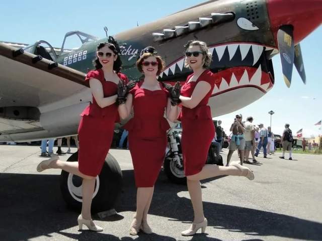 Three women dressed in 50s style red dresses with red glasses posting in front of a plan with shark teeth and eyes painted on it.