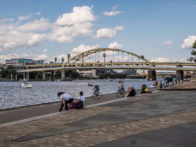 People sitting, biking, and walking around Point State Park in Pittsburgh along a river with a large yellow bridge in the background