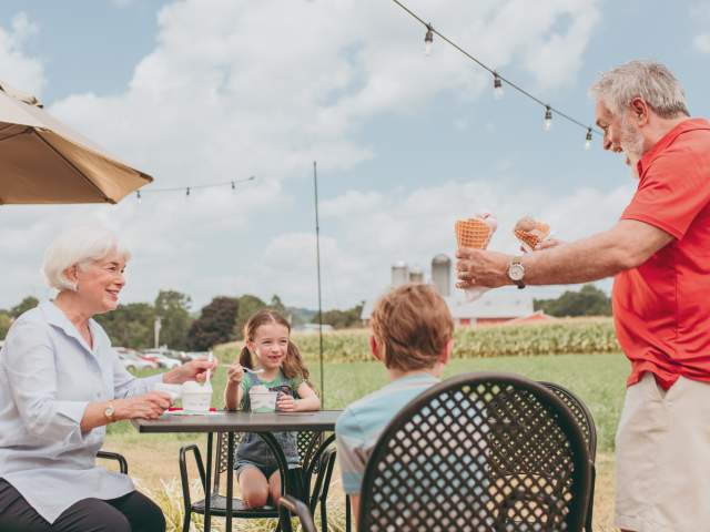 An older man handing two cones of ice cream to an older woman and two children sitting at an outdoor table