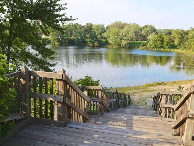 A boardwalk with steps leads down to a lake with trees surrounding it. The water is calm and reflects the trees and sky.