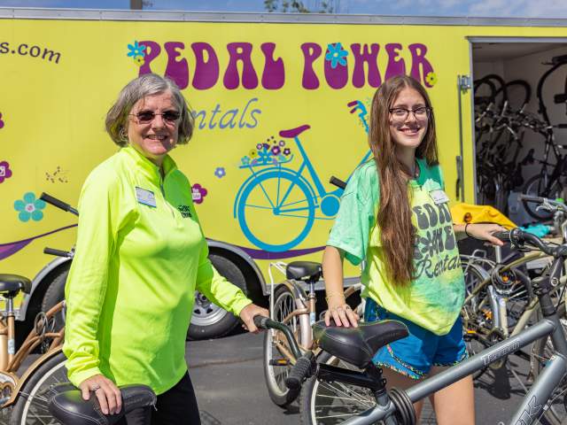 Two women in bright green shirts hold bicycles in front of a bicycle rental trailer.