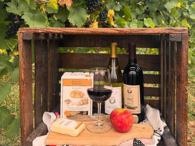 Wooden crate with autumn wines and charcuterie  board with apples, crackers, and cheese in front of Vineyards at Fero Winery in Lewisburg.