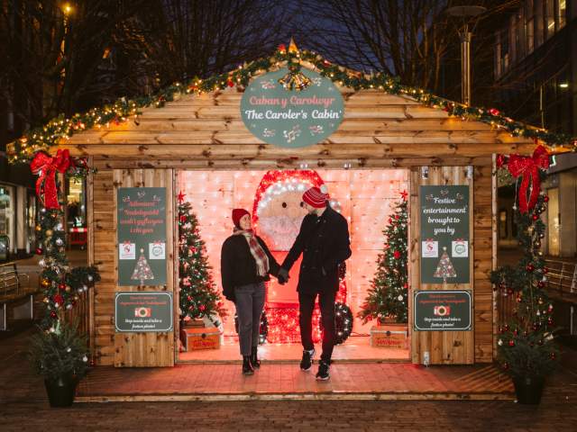 young couple standing in front of a wooden chalet at the swansea christmas market