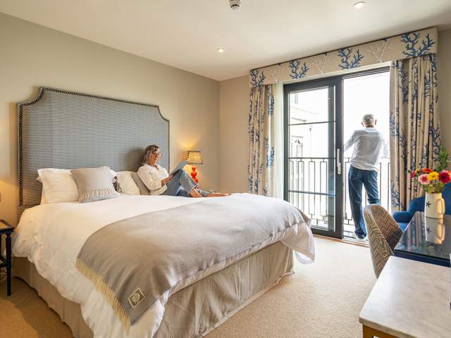 A man and a woman in a hotel room. Woman reading on the bed, man looking out of the window.