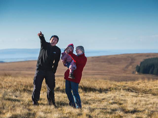 An older couple in winter clothes in a rural landscape, woman is holding a child, the man is pointing to something