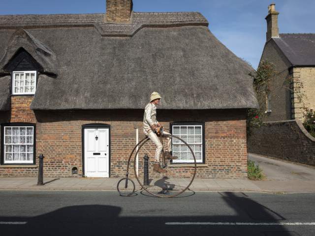 a man on a penny farthing in front of a thatched house
