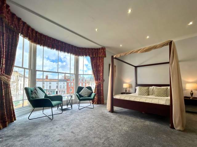 a bed and two chairs in a hotel room by a bay window overlooking a town square