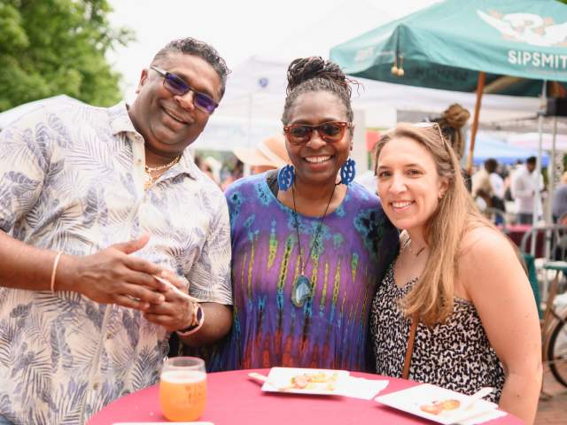 Three people stand at a table smiling at the Wine & Food Festival.