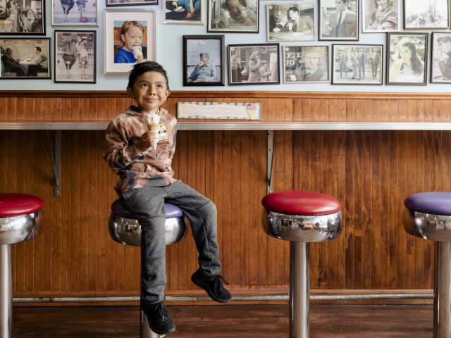 A young boy sits on an ice cream parlor stool with an ice cream cone in his hand.