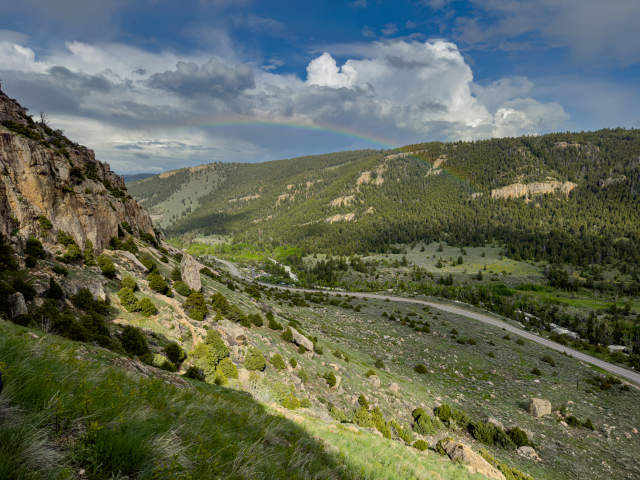 A rainbow glows under the clouds above a lush mountain landscape.