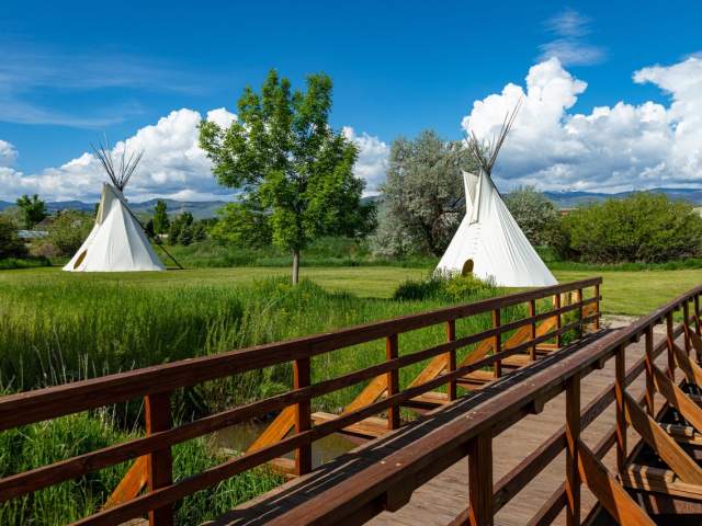 A wooden walkway leads to white teepees standing in Fremont County grasses, with mountains and clouds rising in the distance.