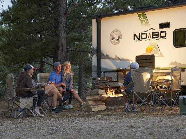 A family gathers in camping chairs around a glowing campfire next to an RV parked in a pine forest during the golden hour.