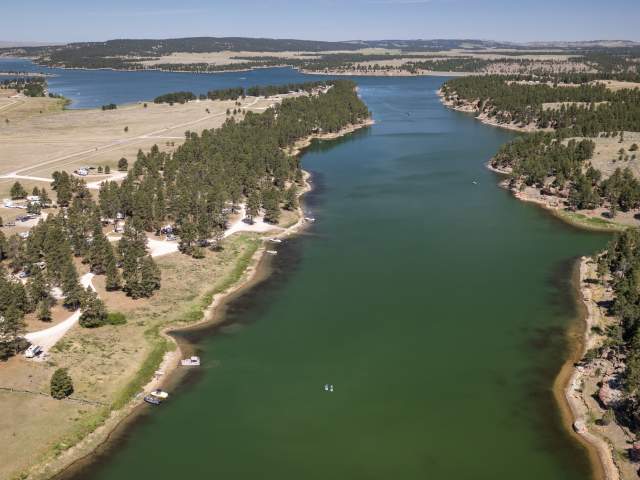 An aerial view shows the winding, teal waters of Keyhole Reservoir framed by pine-covered shorelines and a busy campground dotted with white RVs.