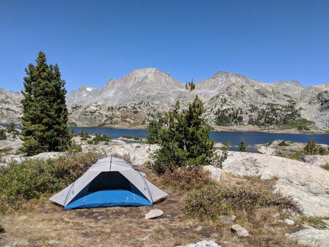 A blue and grey dome tent is pitched on a rocky, alpine ridge overlooking a deep blue mountain lake and jagged granite peaks under a clear sky.