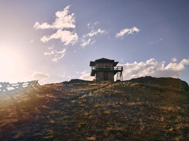 A high-angle, wide shot captures of Sheep Mountain Fire Tower lookout perched on a grassy ridge in Wyoming under a bright, late-afternoon sun.