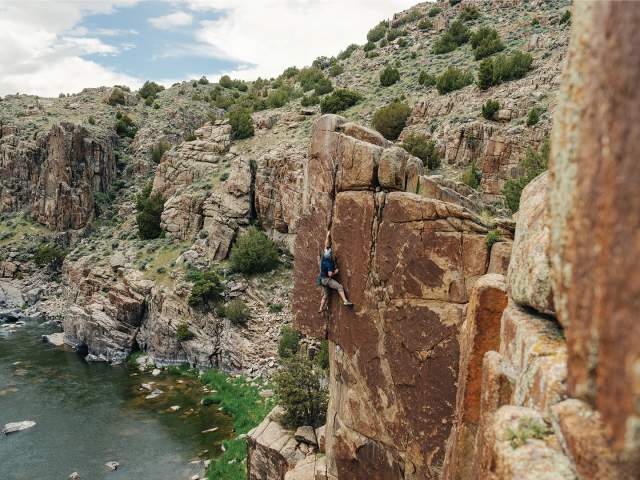A person rock climbs a tall canyon wall above a winding river, surrounded by rugged cliffs and scattered vegetation.