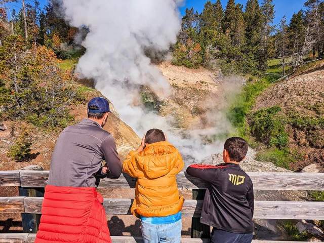 A father and two kids watch as steam erupts from a geyser.  