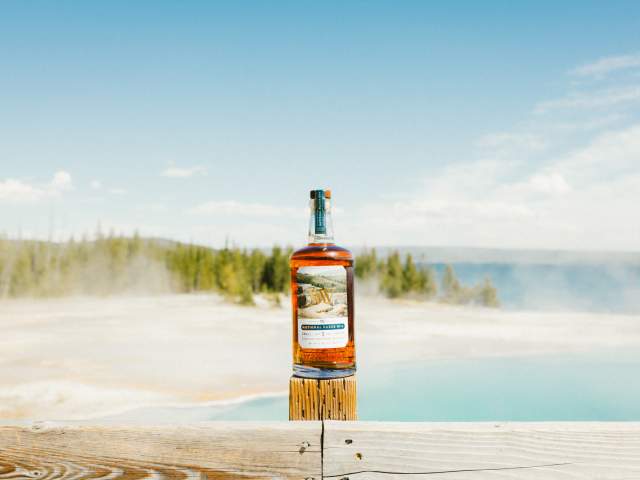 A bottle of Wyoming Whiskey’s National Park Series No. 4 stands on a wooden railing pole in Wyoming with a steaming lake and pine forest as the backdrop.